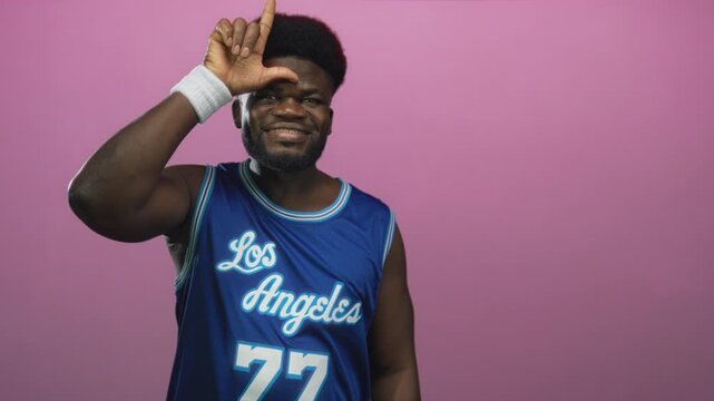 Man wearing blue basketball jersey smiling forms l with hand on forehead in studio against pink wall; playful confidence.