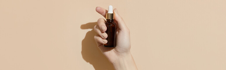 A womans hand holding a brown bottle of serum on a beige background. Body care 