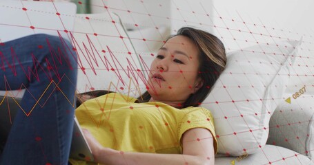 Reclining mid adult Korean woman using tablet on gray sofa, wearing yellow top, jeans, red grid
