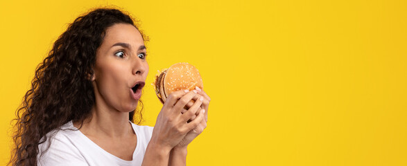 A woman with curly hair expresses excitement while holding a large hamburger in her hands. The vibrant yellow background enhances her joyful reaction.