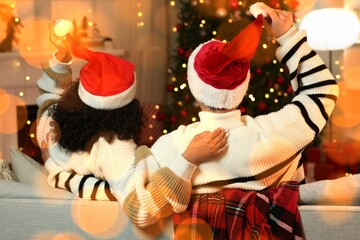 Young couple in warm sweaters and Santa hats sitting on sofa at home on Christmas eve, back view