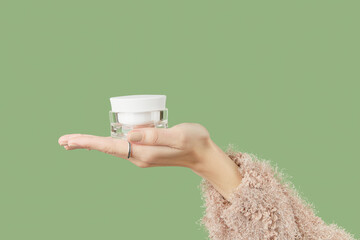 A womans hand in a sweater holds a jar of cream on a green background. Self care beauty treatment