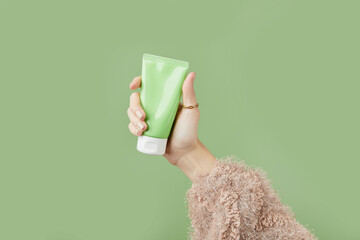A womans hand in a sweater holds a tube of cream on a green background. Self care beauty treatment