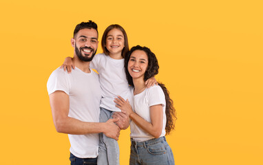 A cheerful family stands together with the father holding their daughter. The mother smiles beside them, all dressed in casual white shirts. The backdrop features a vibrant yellow color.