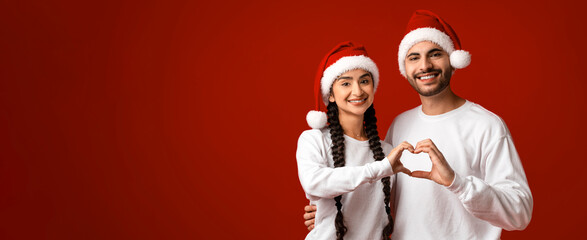 A young couple stands against a bright red background, wearing Santa hats. They are smiling and forming a heart shape with their hands, celebrating the Christmas spirit together.
