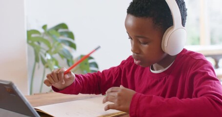 African American boy tapping tablet for school prompts and writing in notebook at home dining table - Powered by Adobe