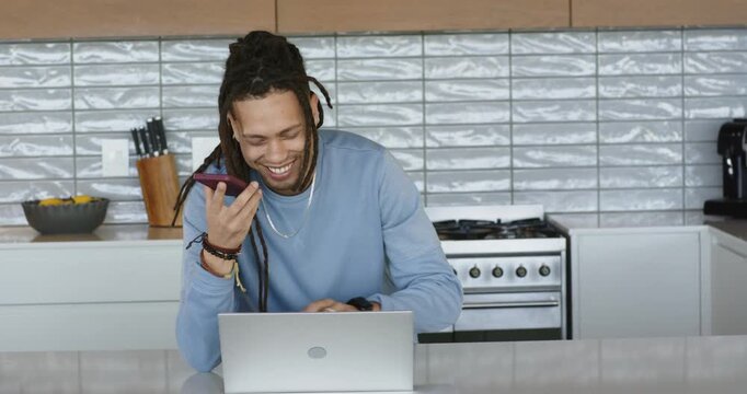 Mid adult man tapping smartphone to start voice call, smiling and leaning toward laptop in kitchen