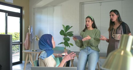 Diverse female coworkers gathering at desk scrolling tablet, tapping laptop, revealing blueprint