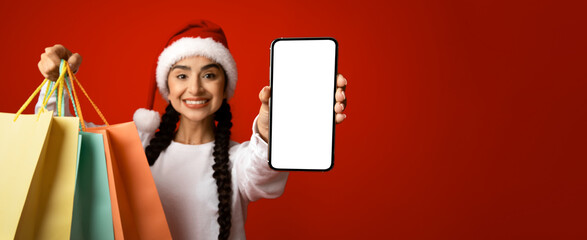 A happy woman wearing a Santa hat smiles widely as she holds colorful shopping bags in one hand and a smartphone displaying a blank screen in the other.
