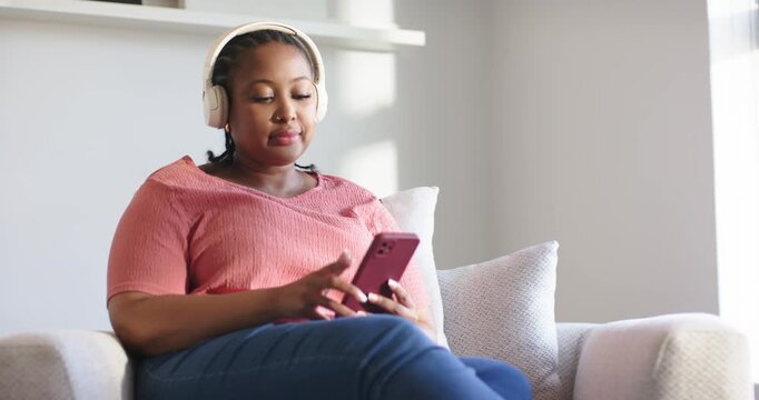 African American woman on sofa wearing headphones lifting smartphone tapping and glancing outside