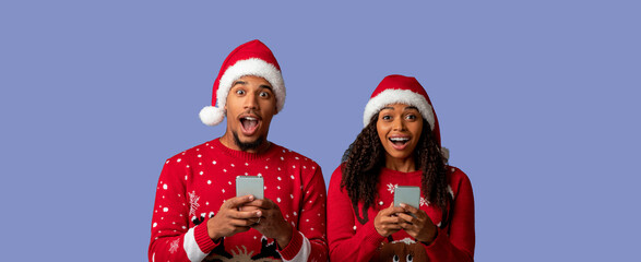 A joyful couple wearing matching Christmas sweaters and Santa hats share an exciting moment while looking at their smartphones.