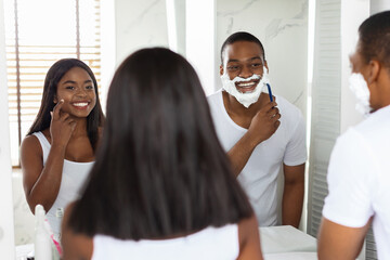 A man shaves in front of a mirror, applying cream on his face, while a woman smiles and gestures nearby, creating a lively morning atmosphere in their bright bathroom.