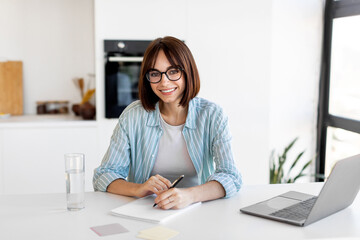 A smiling young woman sits at a stylish desk in a bright room, writing notes in a notepad while a...