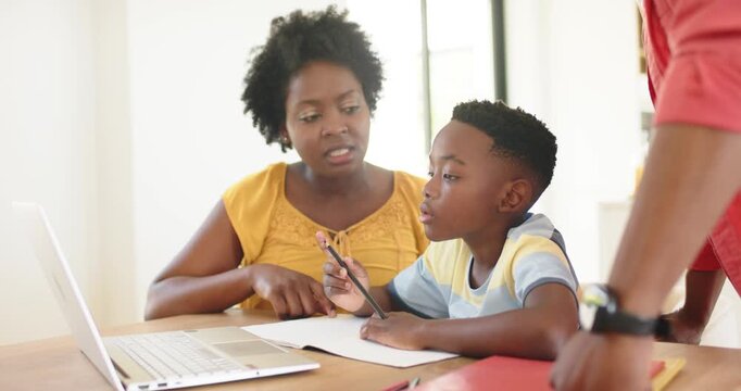 Pointing at laptop screen, African American mother guiding school-age son on homework at home study