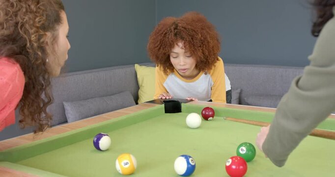 Diverse female friends noticing ball near pocket, blowing it on lounge pool table to sink ball