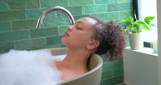 African American woman relaxing in freestanding tub with foam while camera pulling showing faucet