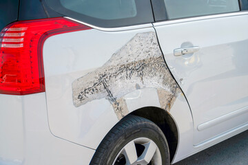 White car with significant body damage on the rear panel, revealing bare metal and filler compound near the wheel well and taillight