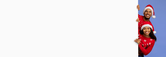 A joyful couple wearing Santa hats and holiday-themed sweaters share a cheerful moment. They are posing against a light blue background, showcasing their festive spirit.