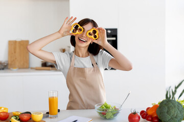 A cheerful woman stands in a bright kitchen holding bell peppers in front of her eyes. She smiles as she prepares a healthy salad, with fresh vegetables and a refreshing drink nearby.
