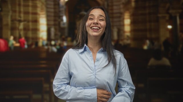 Woman laughing with face toward camera, hands clasped over stomach inside a catholic church nave with wooden pews and stone arches; joy faith.