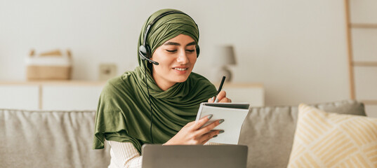 Young woman wearing a green headscarf participates in an online class. She takes notes while...