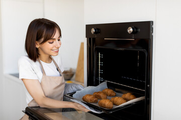 A cheerful woman in an apron reaches into a modern kitchen oven to pull out a tray of golden croissants. Sunlight fills the space, creating a warm atmosphere.
