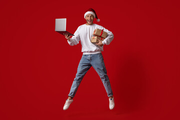 A joyful young man dressed in a Santa hat and festive sweater leaps in the air while holding a...