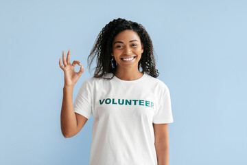 A cheerful volunteer stands in front of a light blue wall, raising her hand to make an okay gesture. She is wearing a white t-shirt that highlights her commitment to helping others.