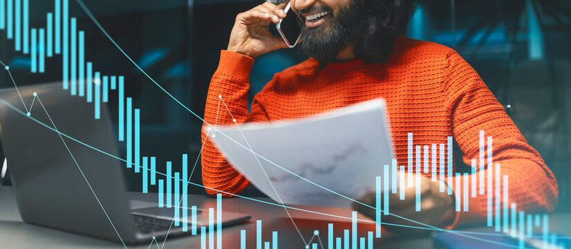 A smiling Indian man in a bright orange sweater engages in a phone call while holding documents. He sits at an office desk with an open laptop during a productive workday. - Powered by Adobe