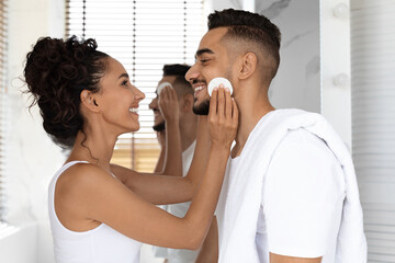 A woman gently applies skincare product to a man's face as they smile at each other. Both are in a...