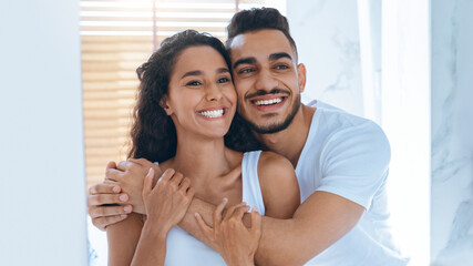 A couple stands closely together, smiling joyfully. They embrace each other in a well-lit room, creating a warm and affectionate atmosphere. Sunlight filters through the window slats.