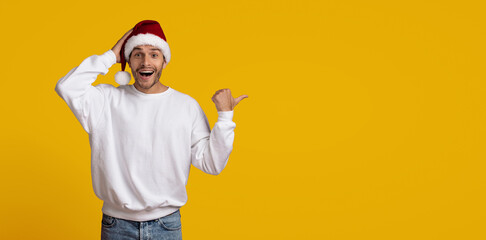A cheerful man wearing a red Christmas hat with white pom-poms smiles and points to the side against a vibrant yellow background. He is dressed in a casual white sweater and jeans.