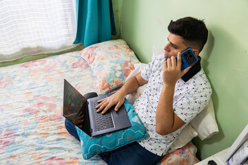 Man focused on laptop while having phone conversation from bed in brightly lit personal bedroom