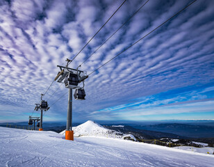 Ski lift chairs moving above a snowy alpine ski resort with skiers on the slopes under a bright blue sky and scattered clouds in a high-altitude mountain landscape