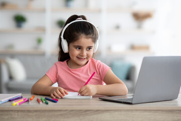 A girl with headphones sits at a desk, smiling as she writes in her notebook during an online learning session. Colorful markers are scattered around her in a cozy living space.