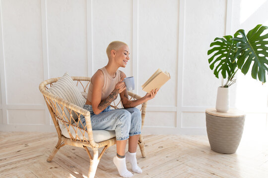 Young woman sitting on wicker chair at home, reading paper book, holding cup and drinking hot coffee. Happy lady taking break from work, copy space - Powered by Adobe