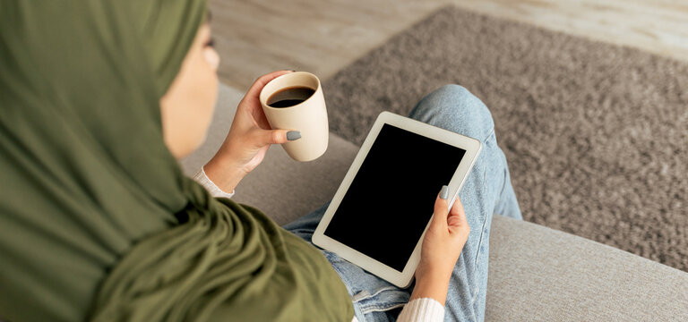A woman with a green headscarf relaxes on a sofa in her living room. She holds a coffee cup in one hand and a tablet in the other, engaged in leisure time within a comfortable interior. - Powered by Adobe
