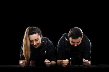 Two fitness enthusiasts in black gear perform plank push ups on a mat, showcasing teamwork, strength, and focus in a dramatic studio setting