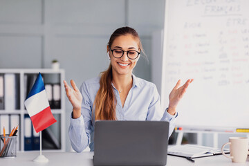 A young woman with glasses is engaging in a video conference. She appears excited as she talks, expressing herself with hand gestures. A small French flag is displayed on her desk.