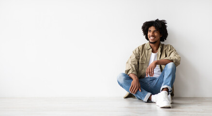 A young African American guy sits on the floor with a relaxed expression, gazing to the side. He appears happy and contemplative, enjoying a moment of peace in a light-filled area.