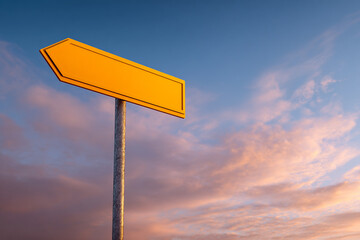 Vibrant yellow directional arrow sign against a beautiful sunset sky with pink and orange clouds.