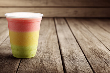 Red, yellow, and green tri-layered smoothie cup on a weathered wooden plank table. Summer healthy drink.