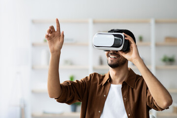 A young man smiles and gestures while wearing a virtual reality headset. He is in a well-lit room with plants and shelves in the background, fully engaged in the virtual experience.
