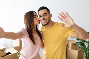 A young couple is smiling in their new home, surrounded by moving boxes. The woman playfully kisses the man while he waves to the camera, showcasing their joy and excitement.