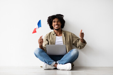 A cheerful black man sits on the floor with his laptop and a small French flag, giving a thumbs up to the camera. He enjoys his distance learning experience in a bright, simple space.