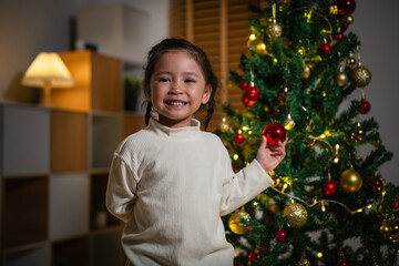 toddler girl decorating toy ball on Christmas tree in home at night