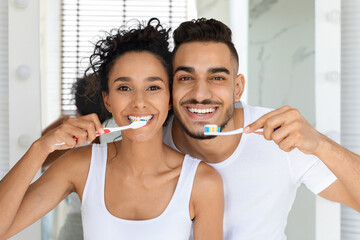 A joyful couple enjoys brushing their teeth in a well-lit bathroom. They smile at each other while using colorful toothbrushes, sharing a fun moment in their daily morning routine.