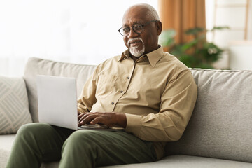 A senior man is focused on his laptop while seated on a light-colored sofa in his living room. Sunlight is streaming in through the windows, creating a warm atmosphere.
