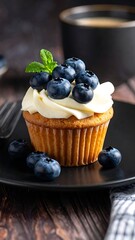 Blueberry cupcake with creamy frosting on a black plate with a dark background and a hint of coffee