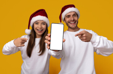 A joyful couple wearing red Christmas hats smiles while pointing at a blank smartphone. The bright...